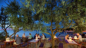 Un espace de restauration en plein air, avec des tables et des bancs en bois, entouré d’oliviers et offrant une vue sur une étendue d’eau.