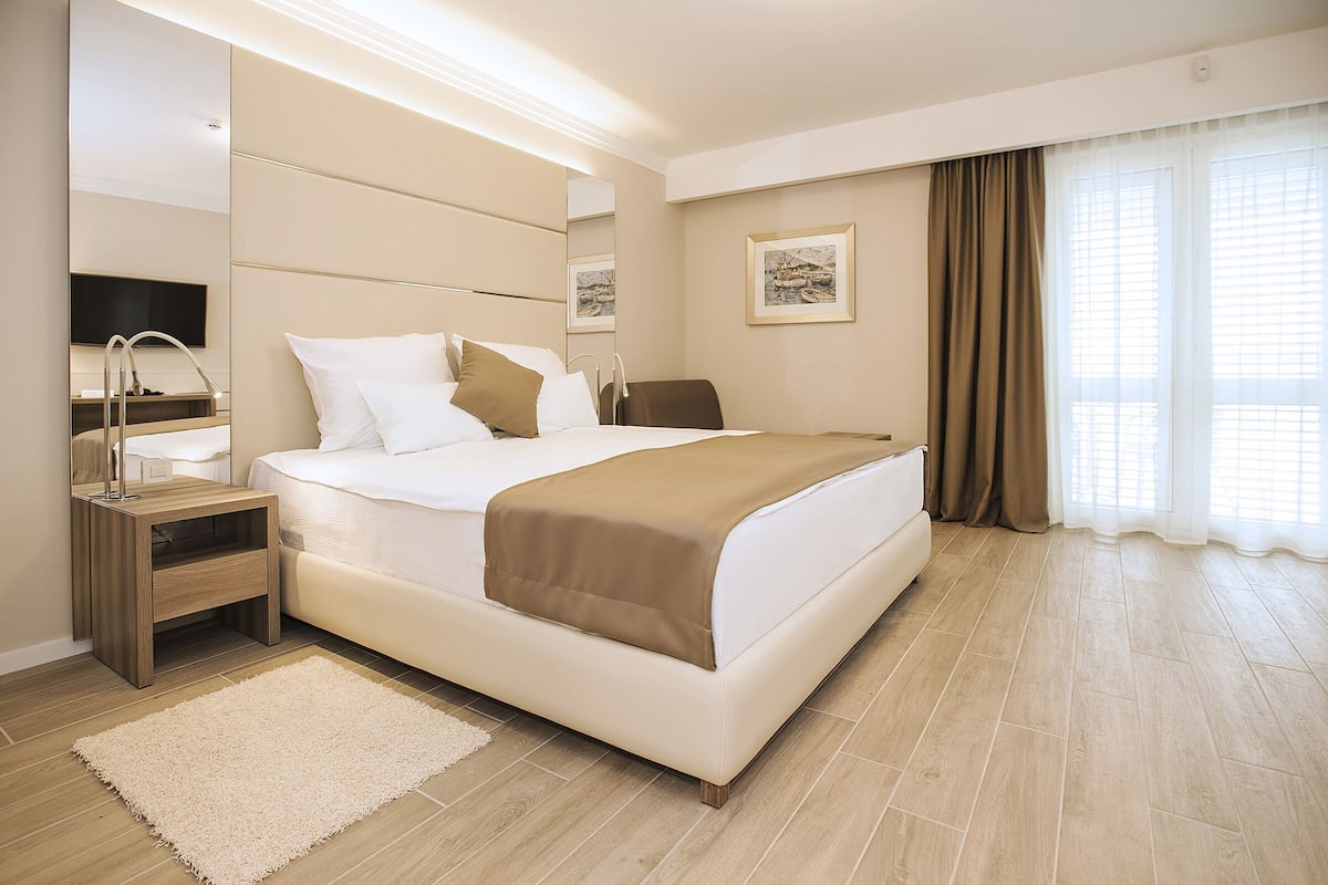 Een moderne hotelkamer met een groot bed, nachtkastje, spiegel, flatscreen-tv en raam met gordijnen.