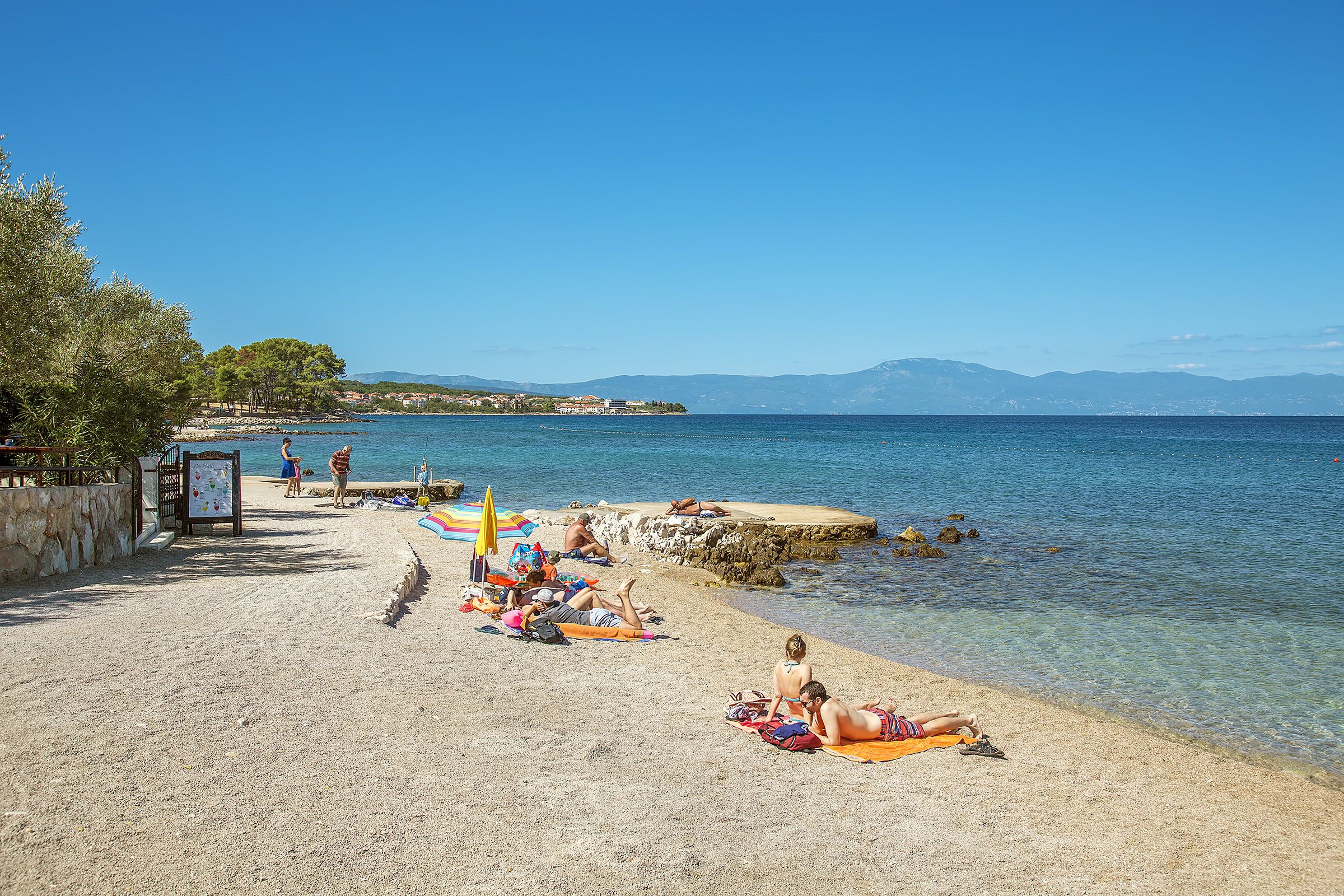 A beach with clear water, people sunbathing, and a distant mountain range.