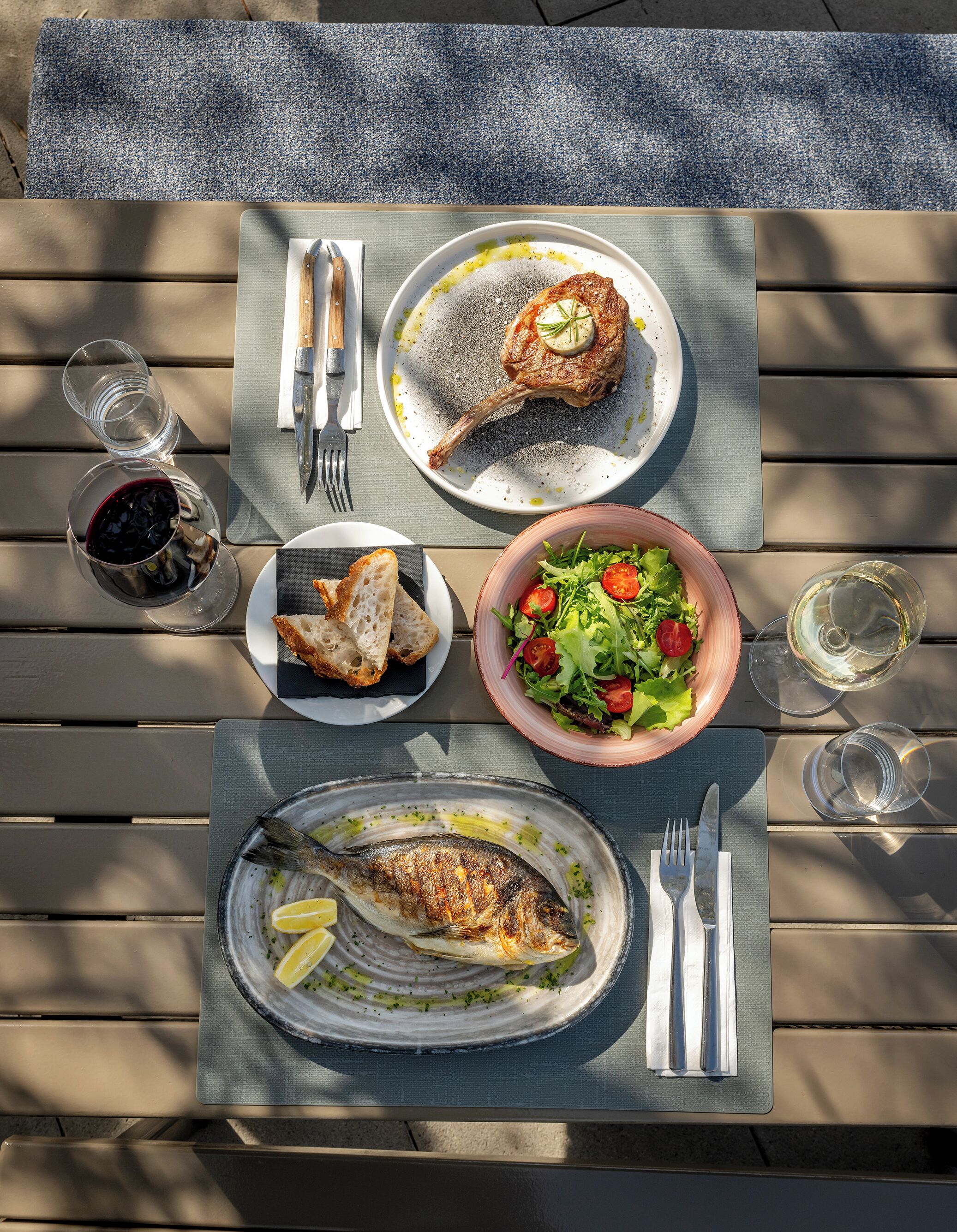 Una mesa servida con un plato de pescado a la parrilla, pan, ensalada y una copa de vino blanco.