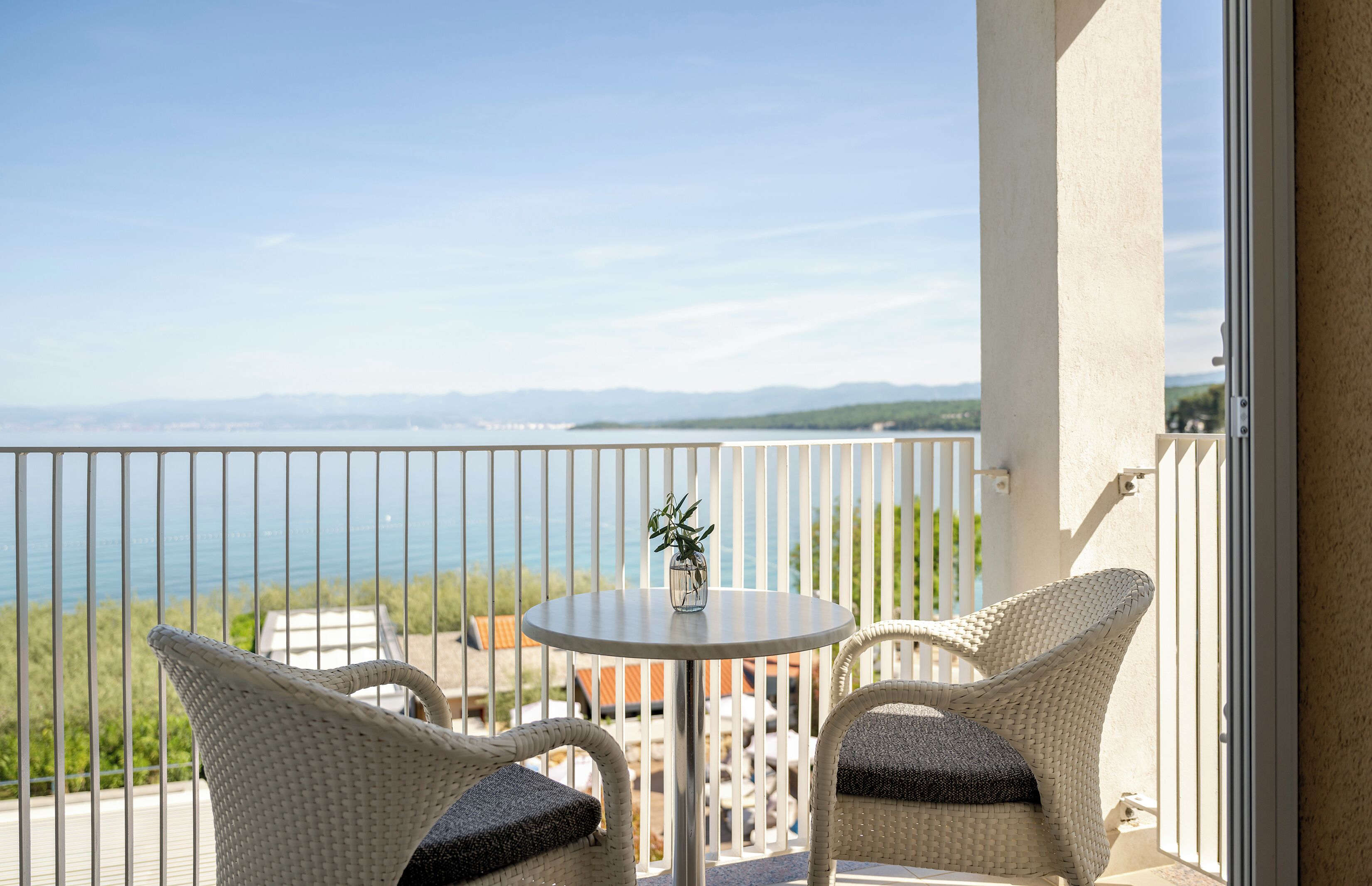 Un balcón con vista al mar, dos sillas de mimbre, una mesita con un florero y una barandilla blanca.