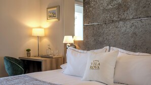 Une chambre d’hôtel avec un lit, deux oreillers ornés du texte « VILA ROVA », un bureau avec une lampe, une chaise et un tableau au mur.