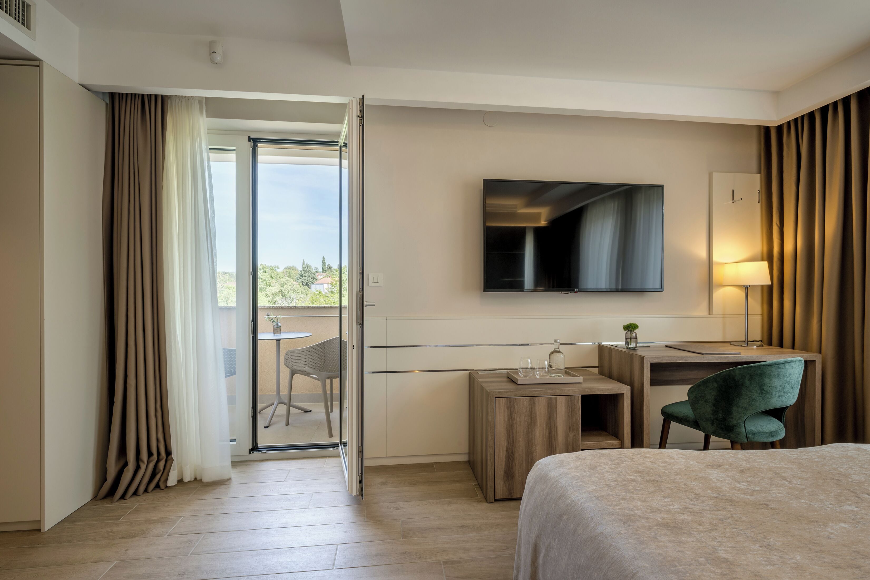 A hotel room with a bed, a desk with a chair, a television, and a sliding glass door leading to a balcony.