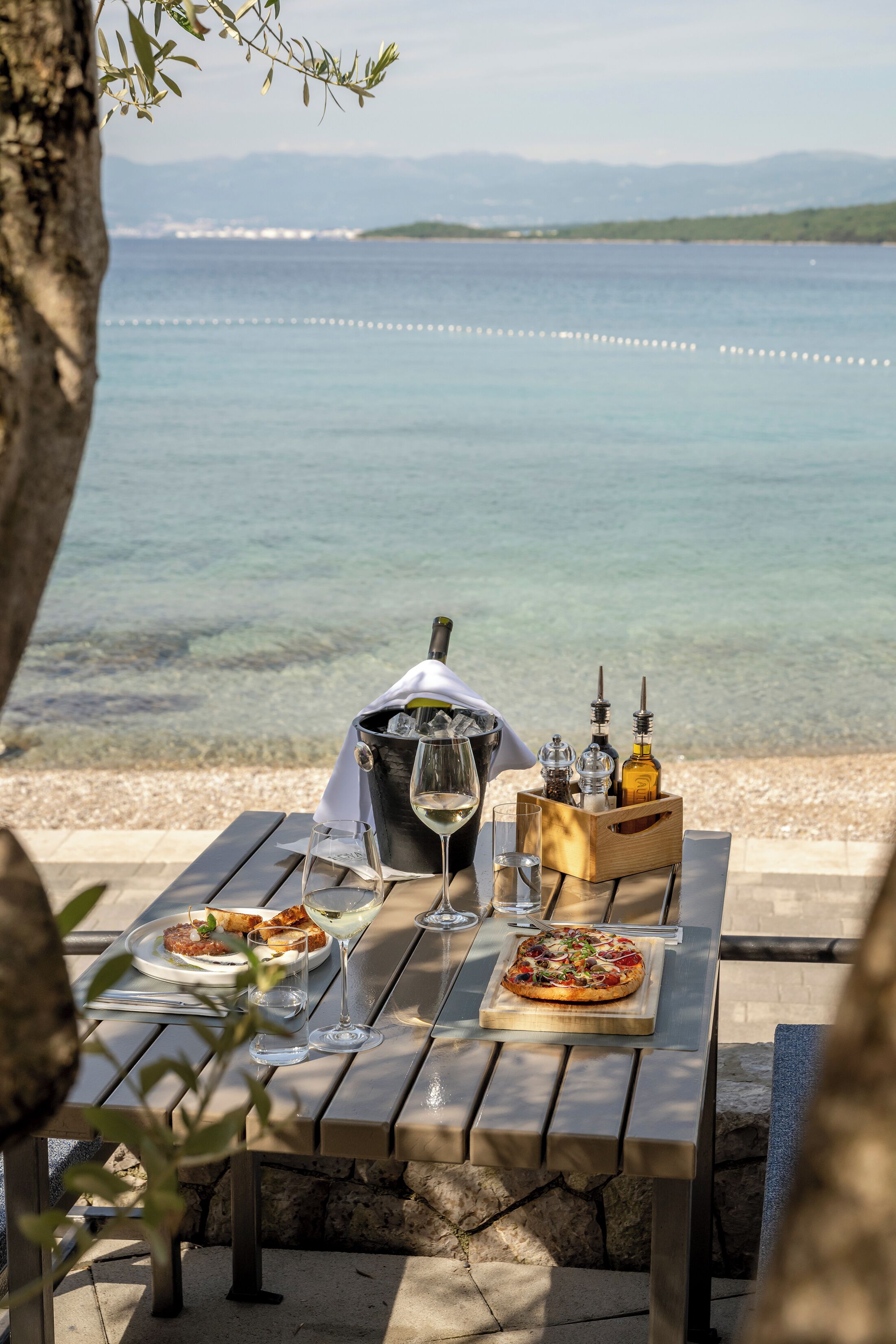 Mesa lista para una comida con vista al mar, una botella de champán y una de aceite de oliva.