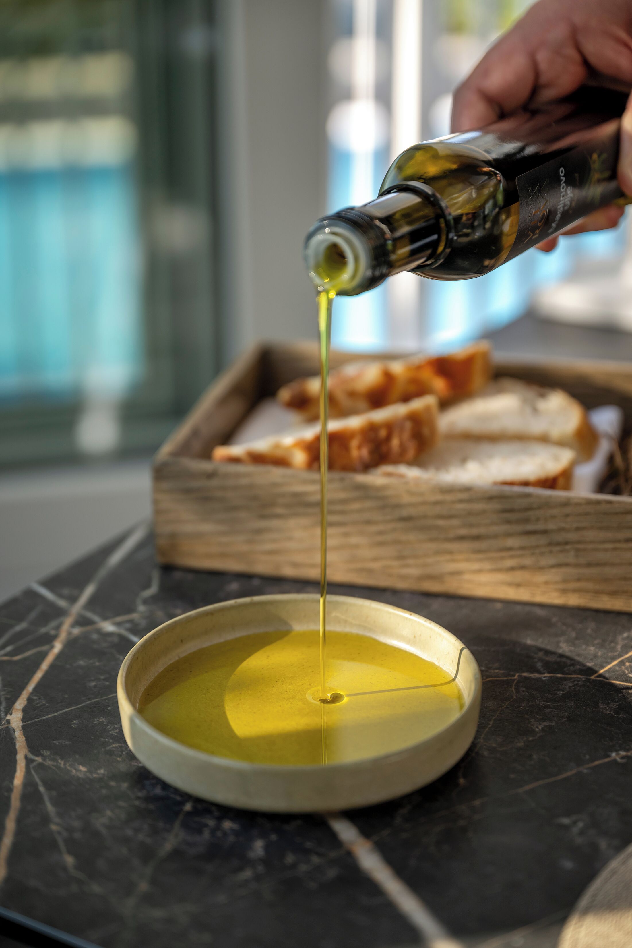 A hand pouring olive oil into a bowl on a marble surface with bread in the background.