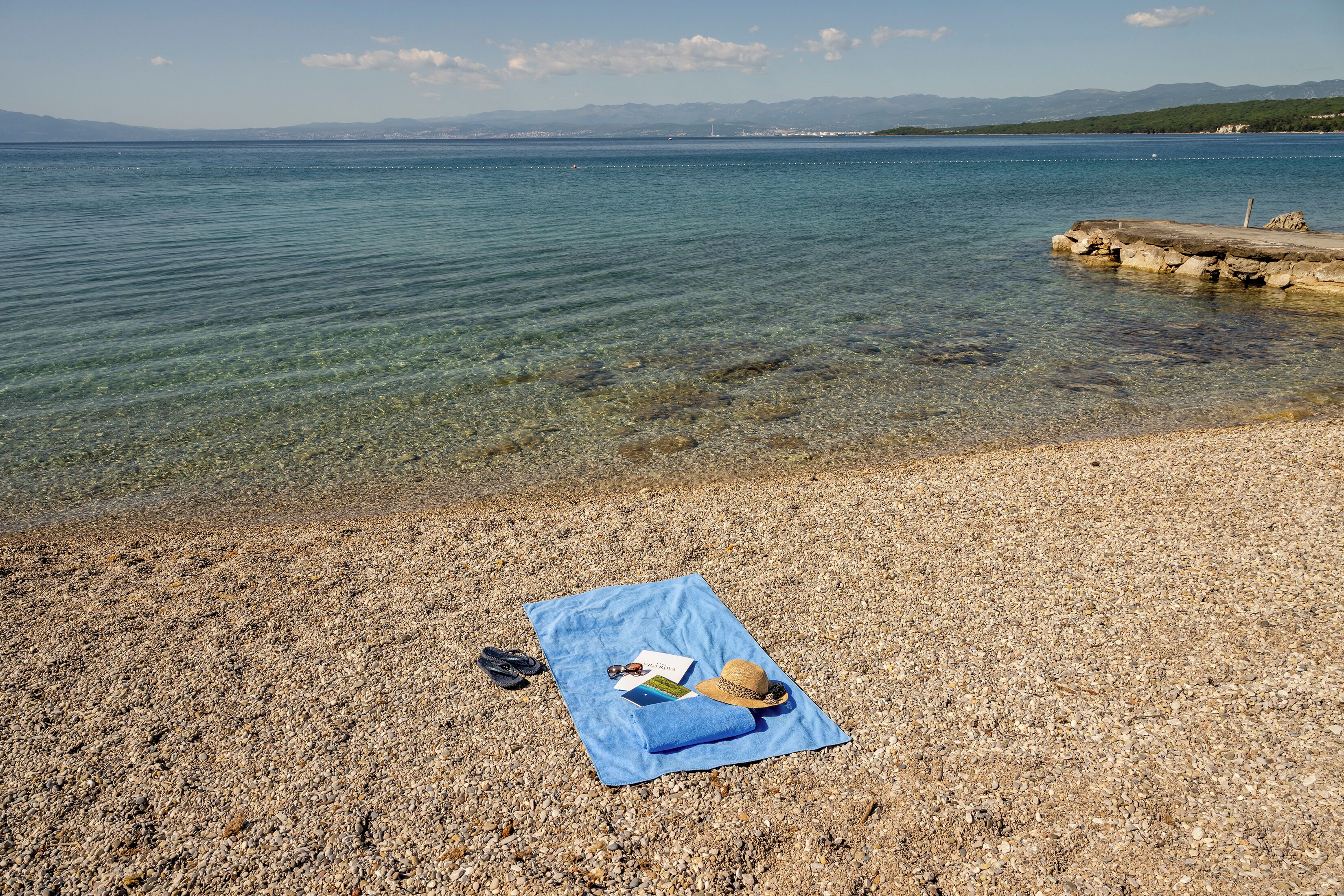 A clear lake with a rocky shore, a blue towel with items on it, and a distant mountain range.