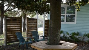 Um banco de madeira e duas cadeiras Adirondack em área externa arborizada, com uma árvore, cerca de madeira e uma casa ao fundo.