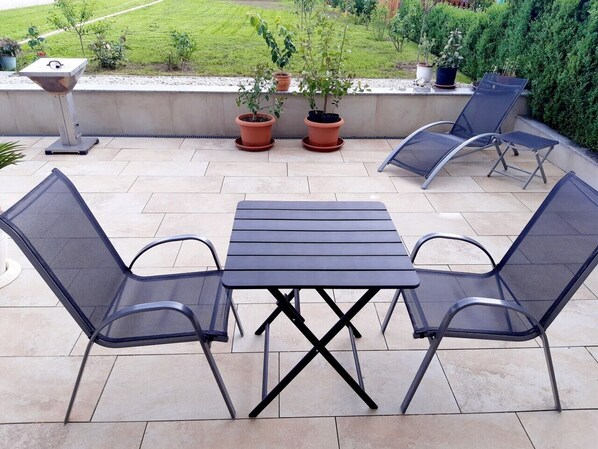 Un patio al aire libre con una mesa, dos sillas, plantas en macetas y una parrilla.