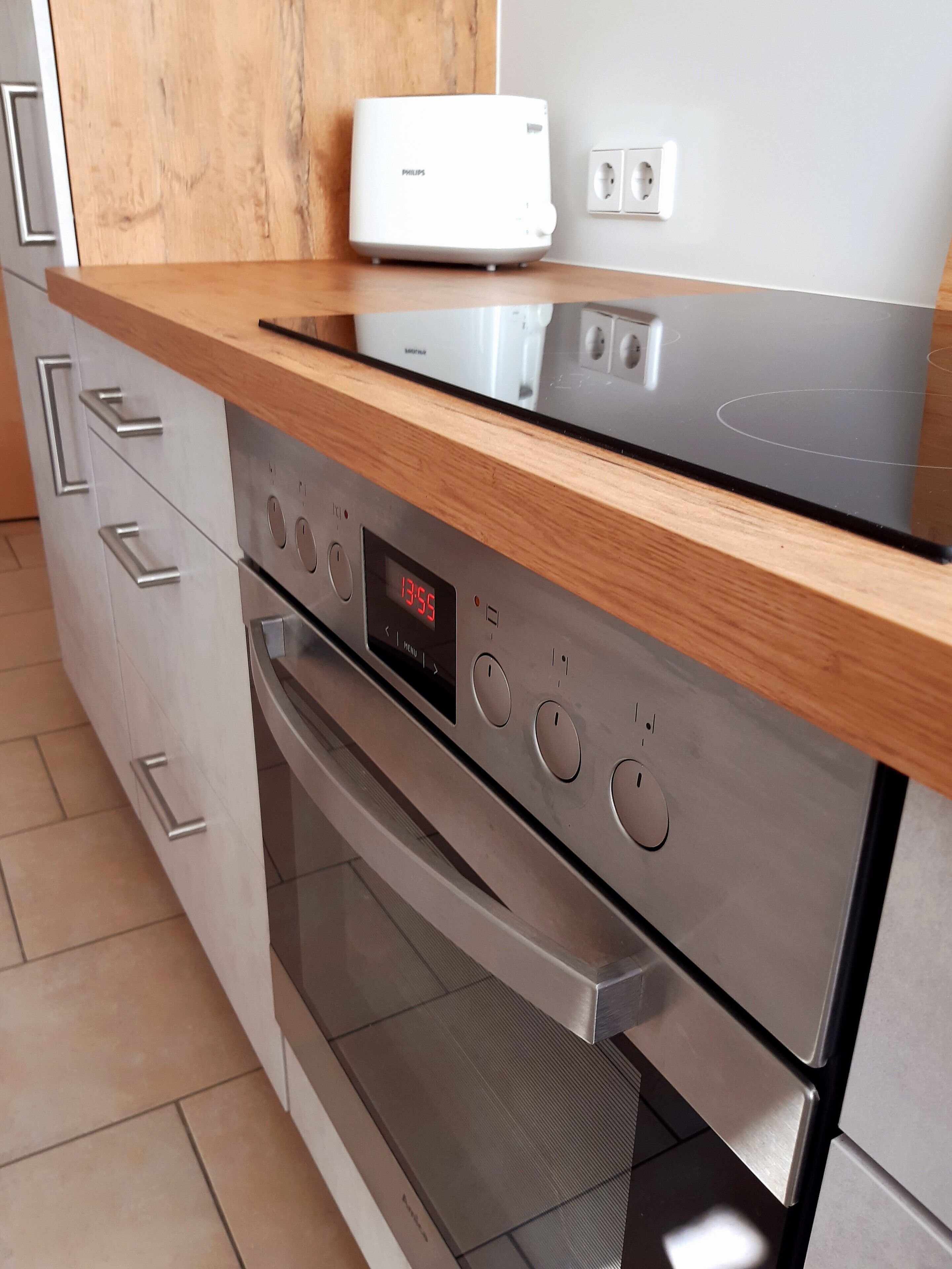 Una cocina moderna con electrodomésticos de acero inoxidable, encimera de madera y un reloj digital que marca las 13:55.