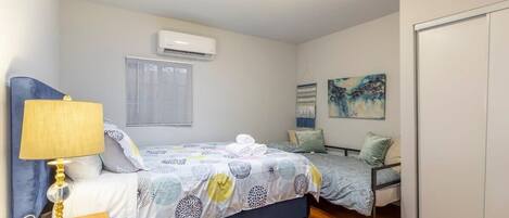 A bedroom with two beds, a ceiling fan, an air conditioning unit, and a painting on the wall.