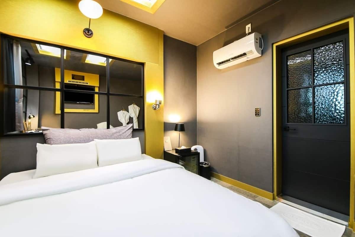 A hotel room with a bed, a mirror, an air conditioning unit, a door, and a window.