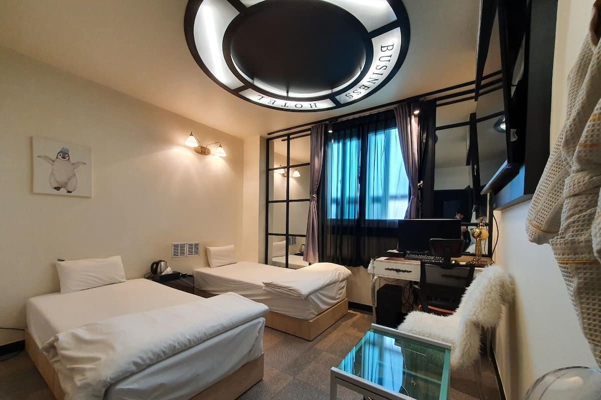 A hotel room with two beds, a desk with a computer, a glass table, and a large window with curtains.