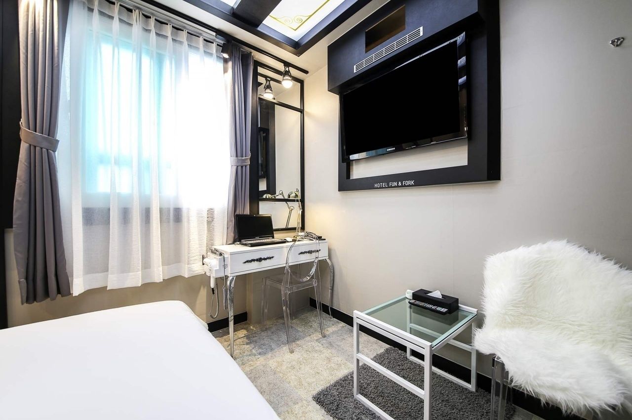 A modern hotel room with a bed, a desk with a laptop, a TV mounted on the wall, and a fluffy chair.