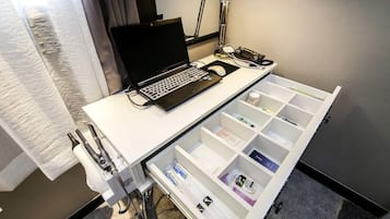 A hotel room with a white desk, a laptop, a hairdryer, and a drawer organizer.