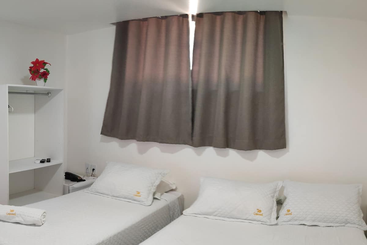 A hotel room with two single beds, a small bedside table, a shelf with toiletries, and a window with brown curtains.