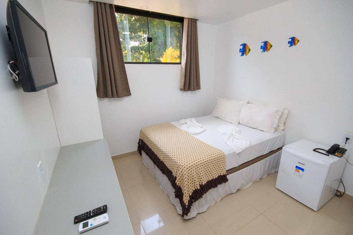 A bedroom with a bed, a bedside table, a TV, a window with curtains, and a small refrigerator.