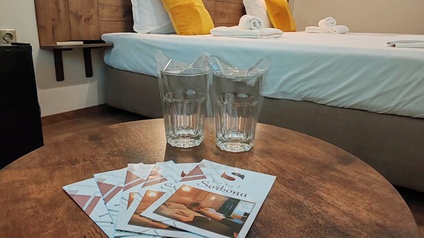 A hotel room with a wooden headboard, a bed with white and yellow pillows, two glasses of water, and a table with brochures. - Hotel Sorbona (Varna)