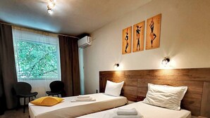 A hotel room with two beds, a window with a view of greenery, a wall-mounted air conditioner, and artwork above the beds. - Hotel Sorbona (Varna)