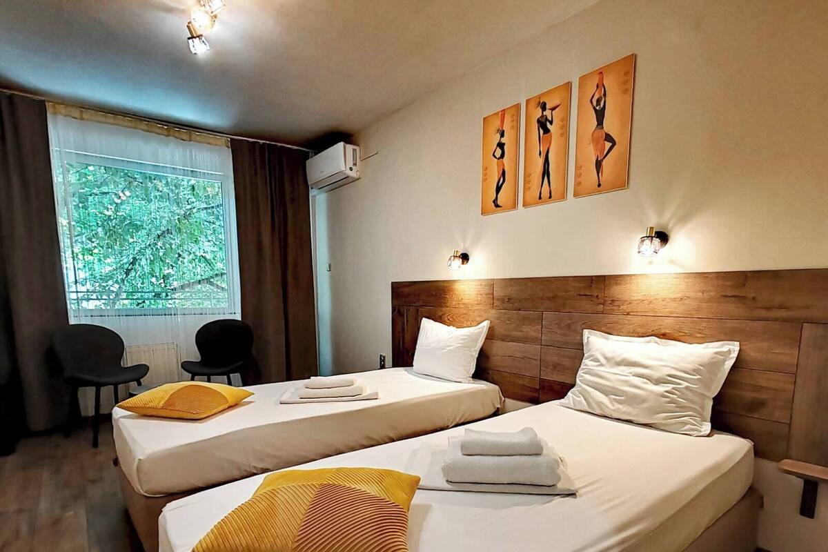 A hotel room with two beds, a window with a view of greenery, a wall-mounted air conditioner, and artwork above the beds.