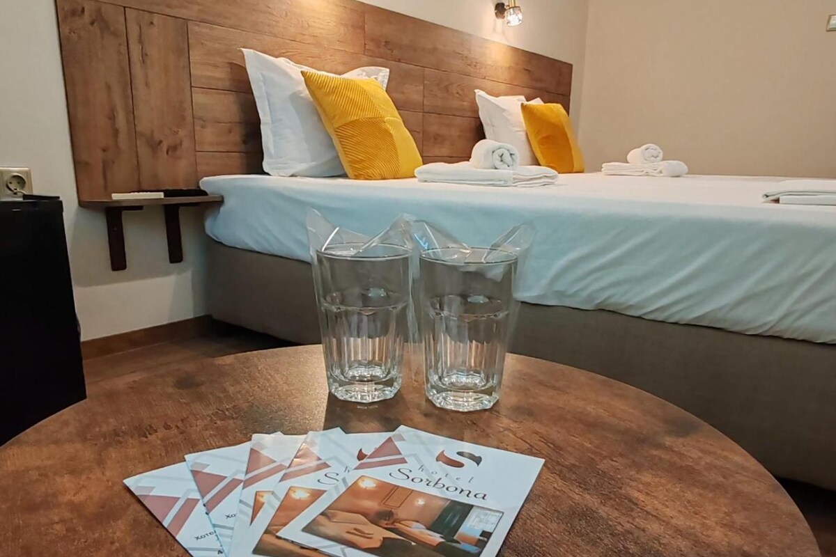 A hotel room with a bed, pillows, a bedside table with glasses of water, and a magazine on the table.