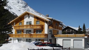 Un hotel de varios pisos con balcones de madera, una montaña nevada al fondo y un área de estacionamiento con automóviles.