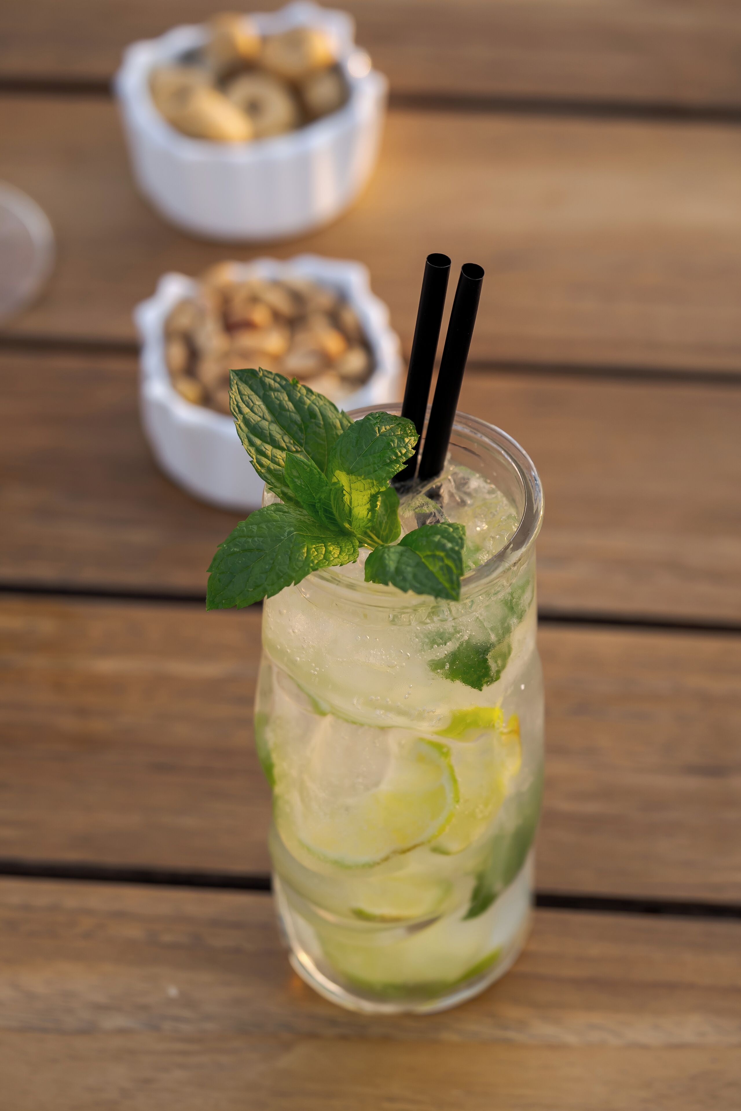 A glass of iced drink with mint leaves and lime slices, with two black straws, on a wooden surface.