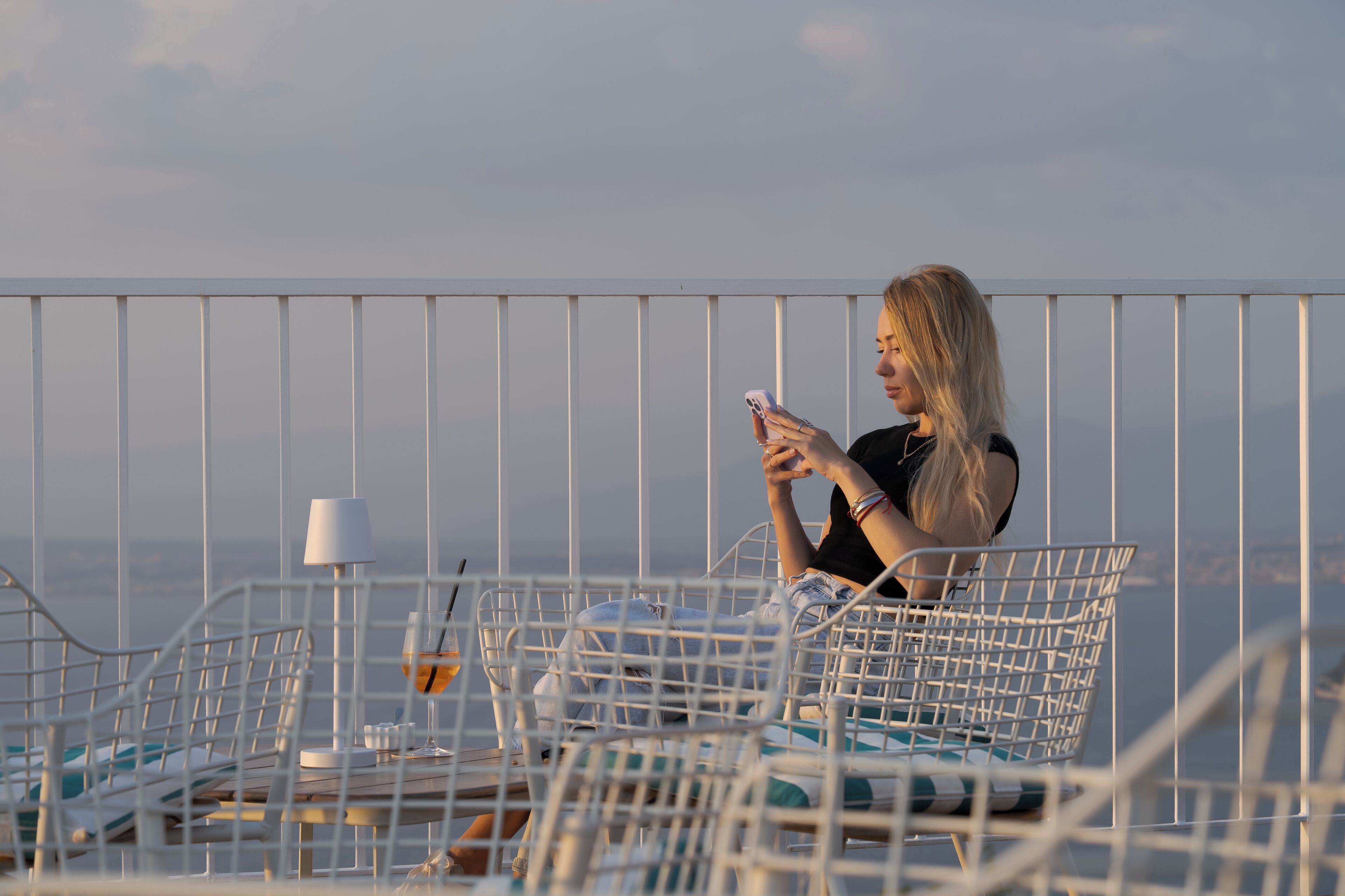 Une personne assise sur une chaise en métal blanche, devant une table, et tenant un téléphone intelligent.