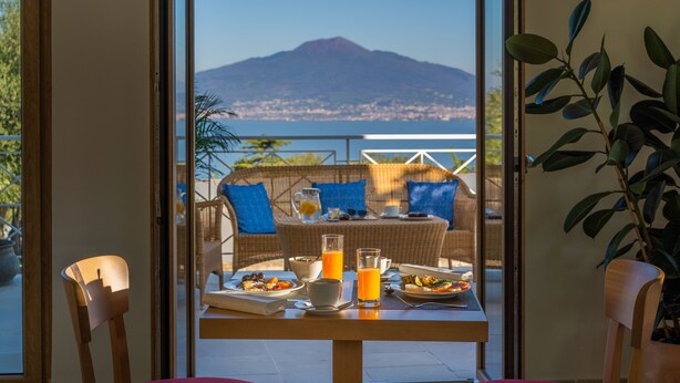 Uma área de refeições com uma mesa posta para o café da manhã, um sofá de vime e vista para o mar e montanha.