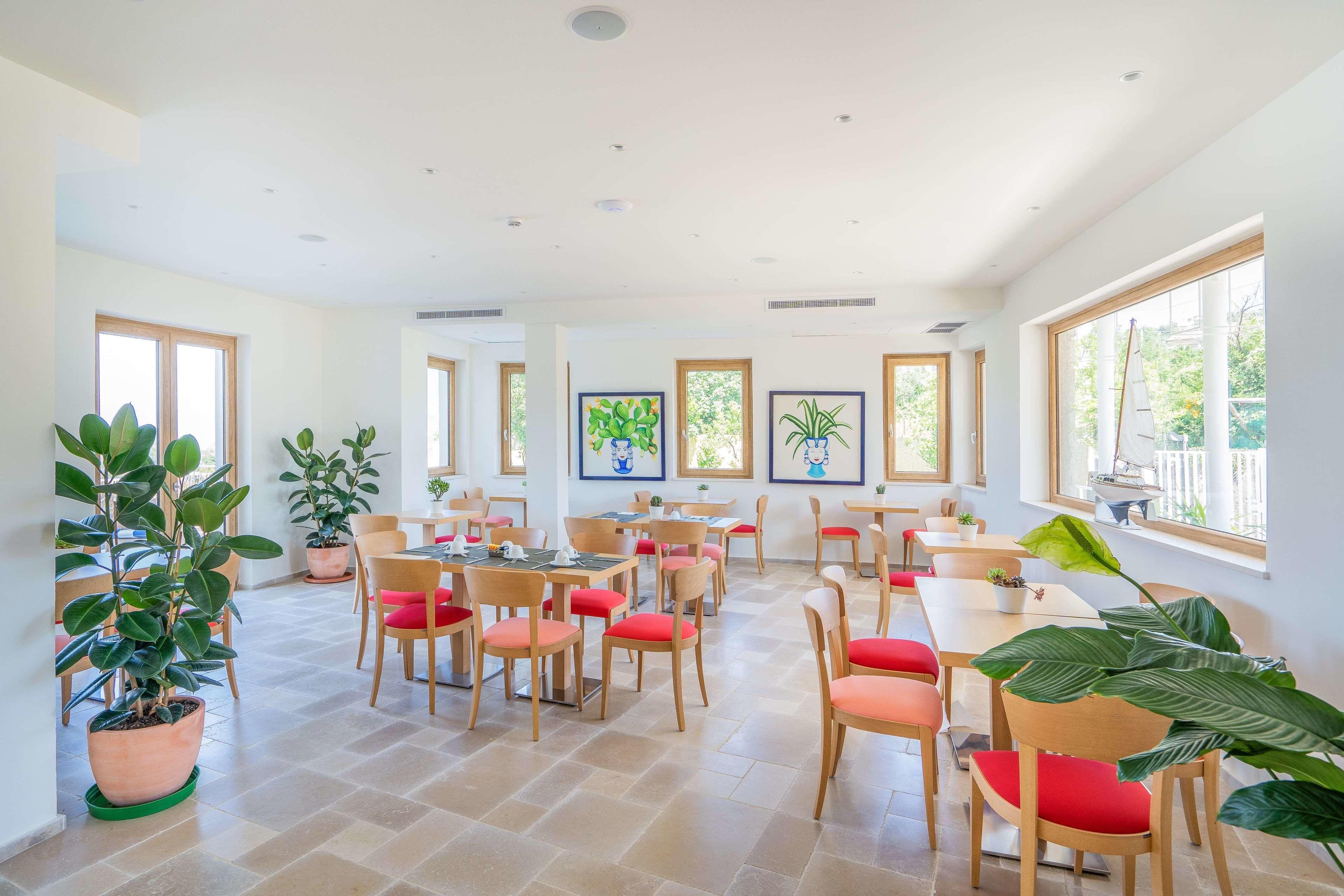 Un comedor amplio con varias mesas y sillas, ventanas grandes y plantas en macetas.