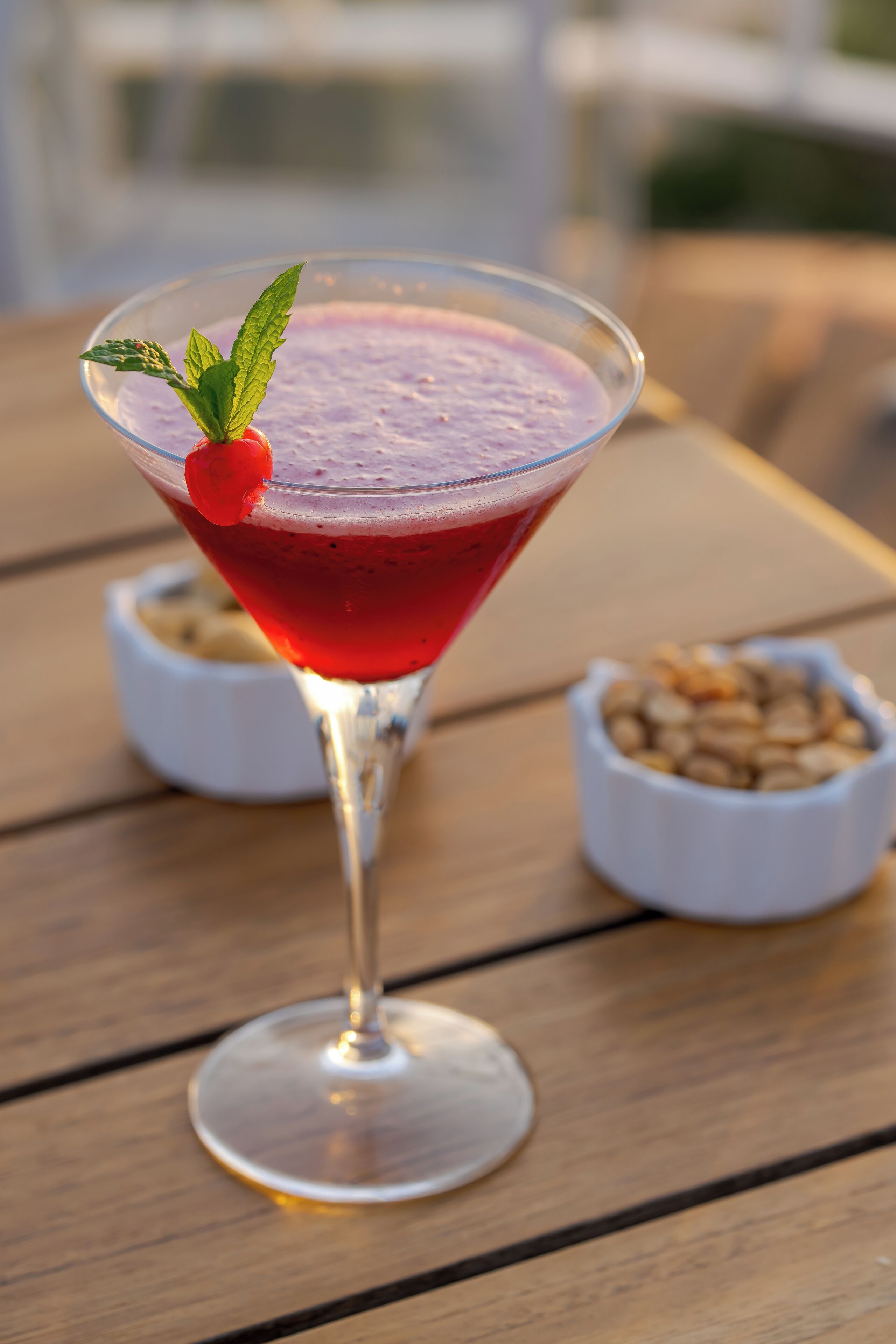 A martini glass with a red drink, garnished with a cherry and mint leaves, on a wooden table with two small bowls in the background.