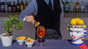 Um barman preparando uma bebida, um vaso com planta e uma cabeça de cerâmica decorativa com limões.