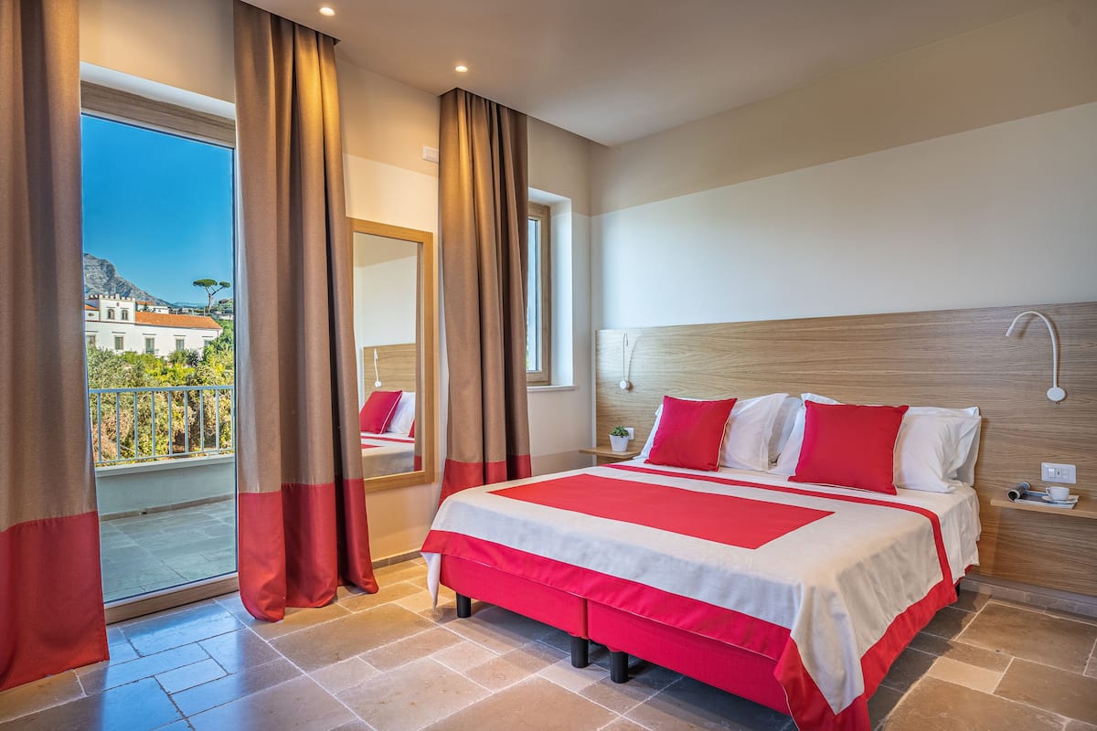 A hotel room with a large bed, a balcony with curtains, a wooden headboard, and a view of the city.