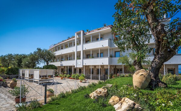 A multi-story hotel with balconies, a garden area with potted plants, and a large tree.