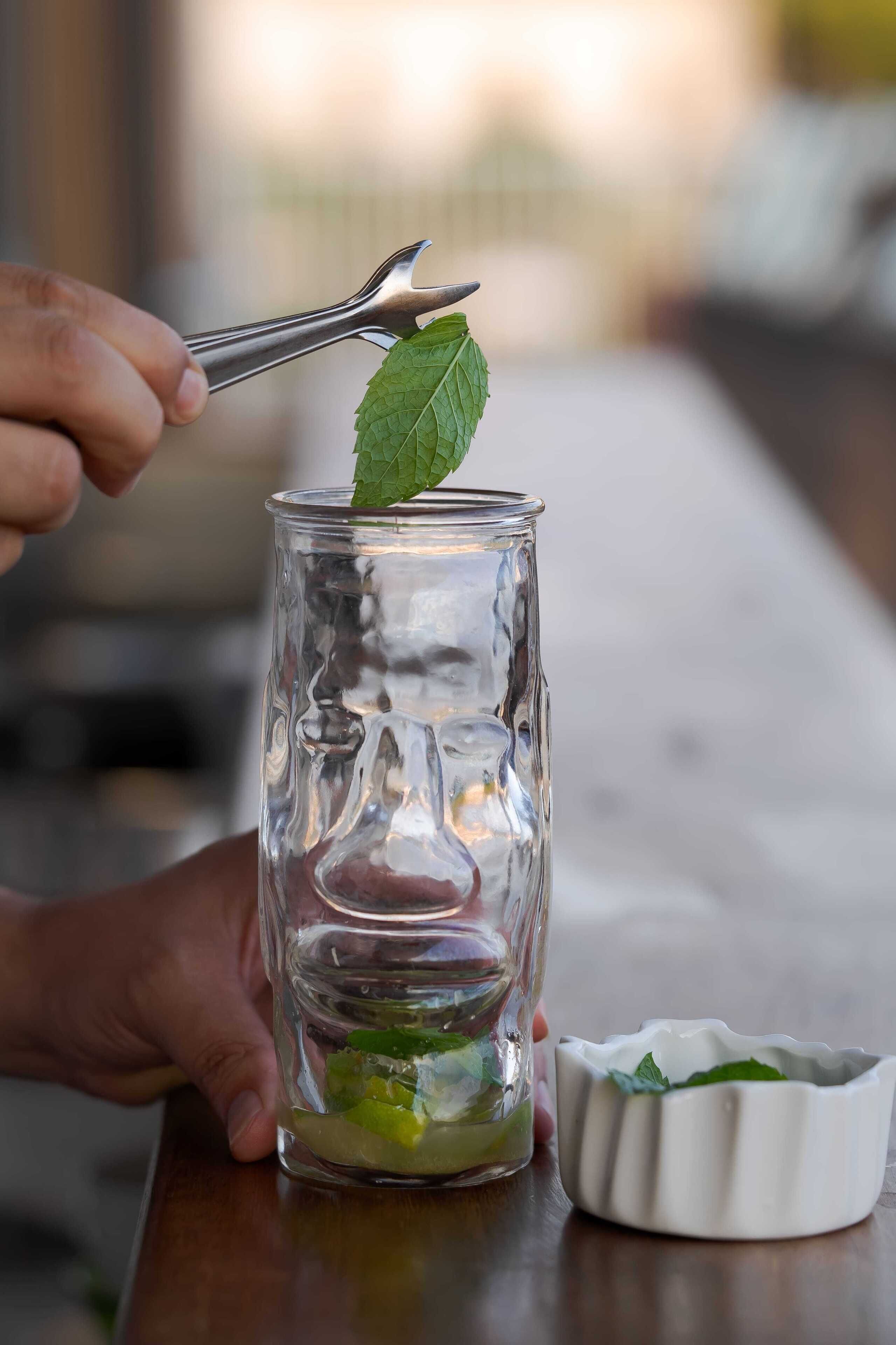 A person holding a pair of tongs over a glass jar containing green leaves and mint, with a small white dish beside it.