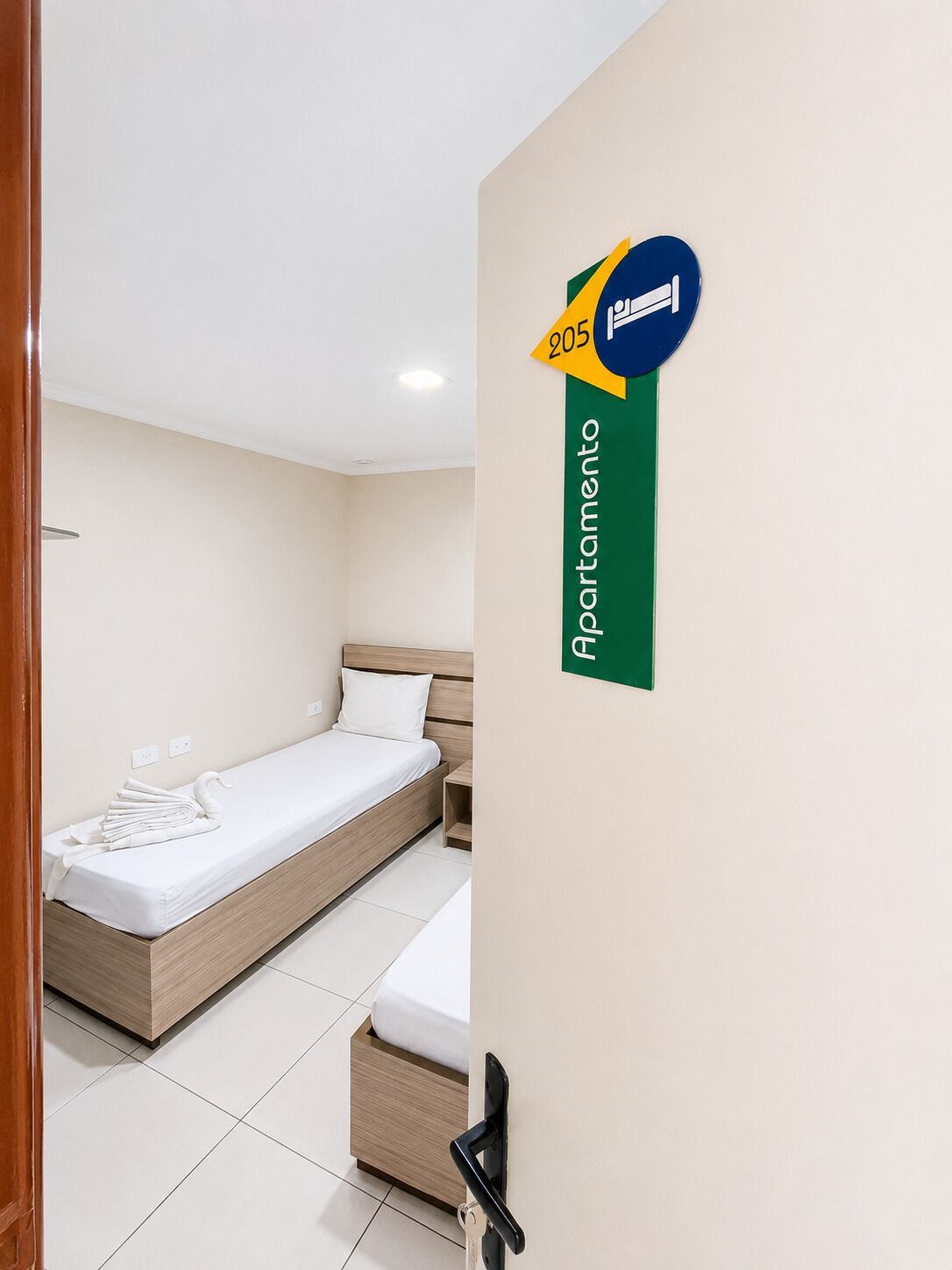 A hotel room with a bed, a small bedside table, a wall-mounted sign indicating room number 205, and a door with a keyhole.