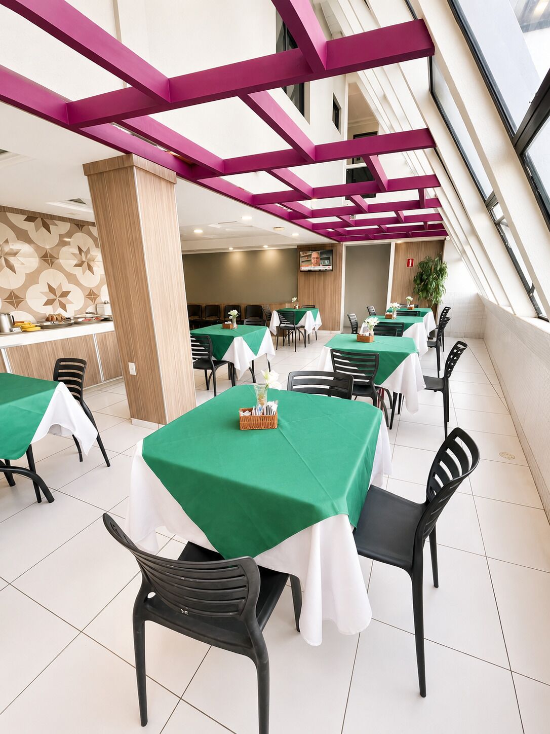 A dining area with tables covered in green tablecloths, black chairs, and a modern ceiling with purple beams.