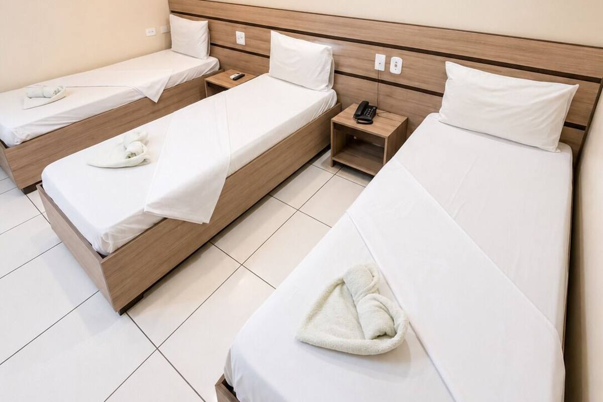 Una habitación de hotel con tres camas individuales, cada una con cabecera de madera, ropa de cama blanca y un control remoto en la mesita de noche.
