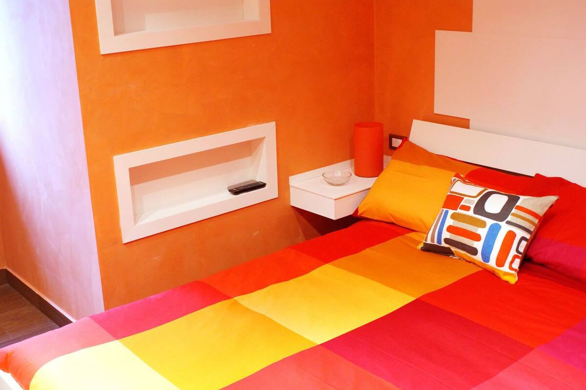 Ein Schlafzimmer mit einem Bett, einem Nachttisch, einer roten Vase und einem farbenfrohen Kissen.