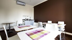 Een moderne eetruimte met witte tafels en stoelen, een kleine kitchenette en een bruine accentmuur.