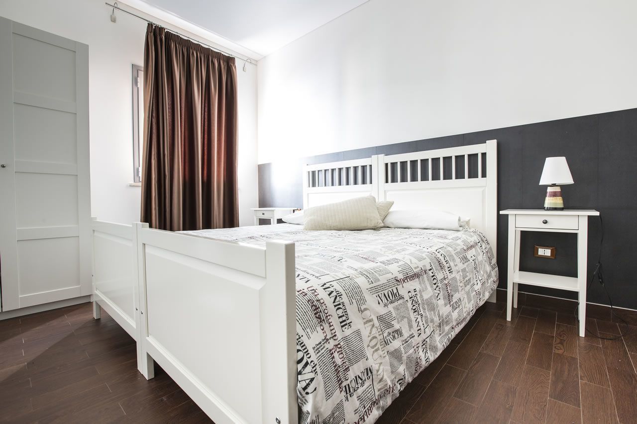 Une chambre à coucher avec un lit blanc, une table de chevet, une lampe et une fenêtre avec des rideaux.