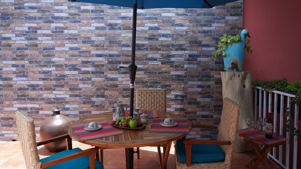 An outdoor patio with a brick wall, a round wooden table, wicker chairs, and a blue umbrella. - Casa Alfareros (Tlaquepaque)