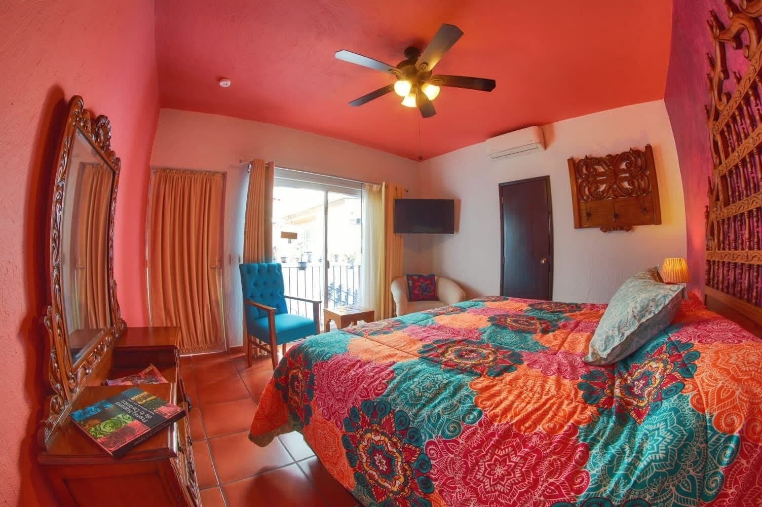 A bedroom with a large bed, a ceiling fan, a TV, a chair, and a balcony with curtains.