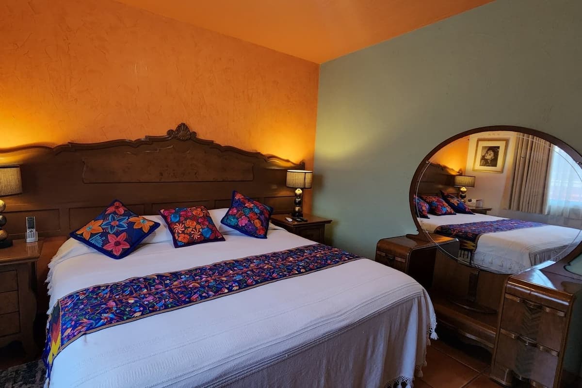 A hotel room with a large bed, a wooden headboard, two bedside tables with lamps, a mirror, and a small bedside table with a book and a sign.