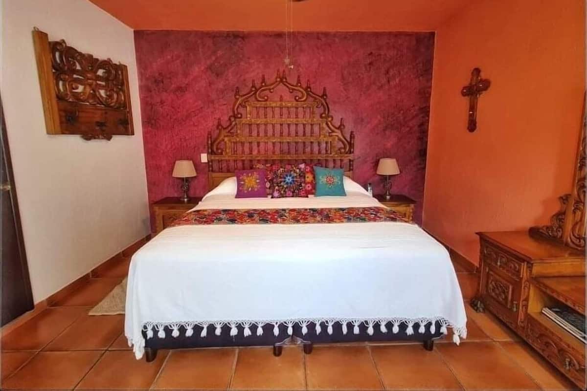 A bedroom with a wooden headboard, a bed with a white bedspread and colorful pillows, two bedside tables with lamps, and a wooden cabinet.