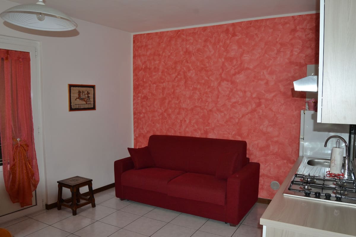 A living room with a red sofa, a small wooden stool, a kitchen area with a gas stove, and a red patterned wall.