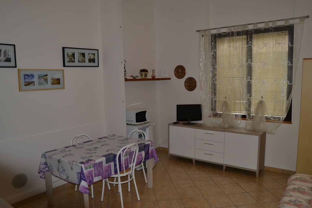 A small, tidy room with a dining table, chairs, a white desk with a computer, and a window with curtains.