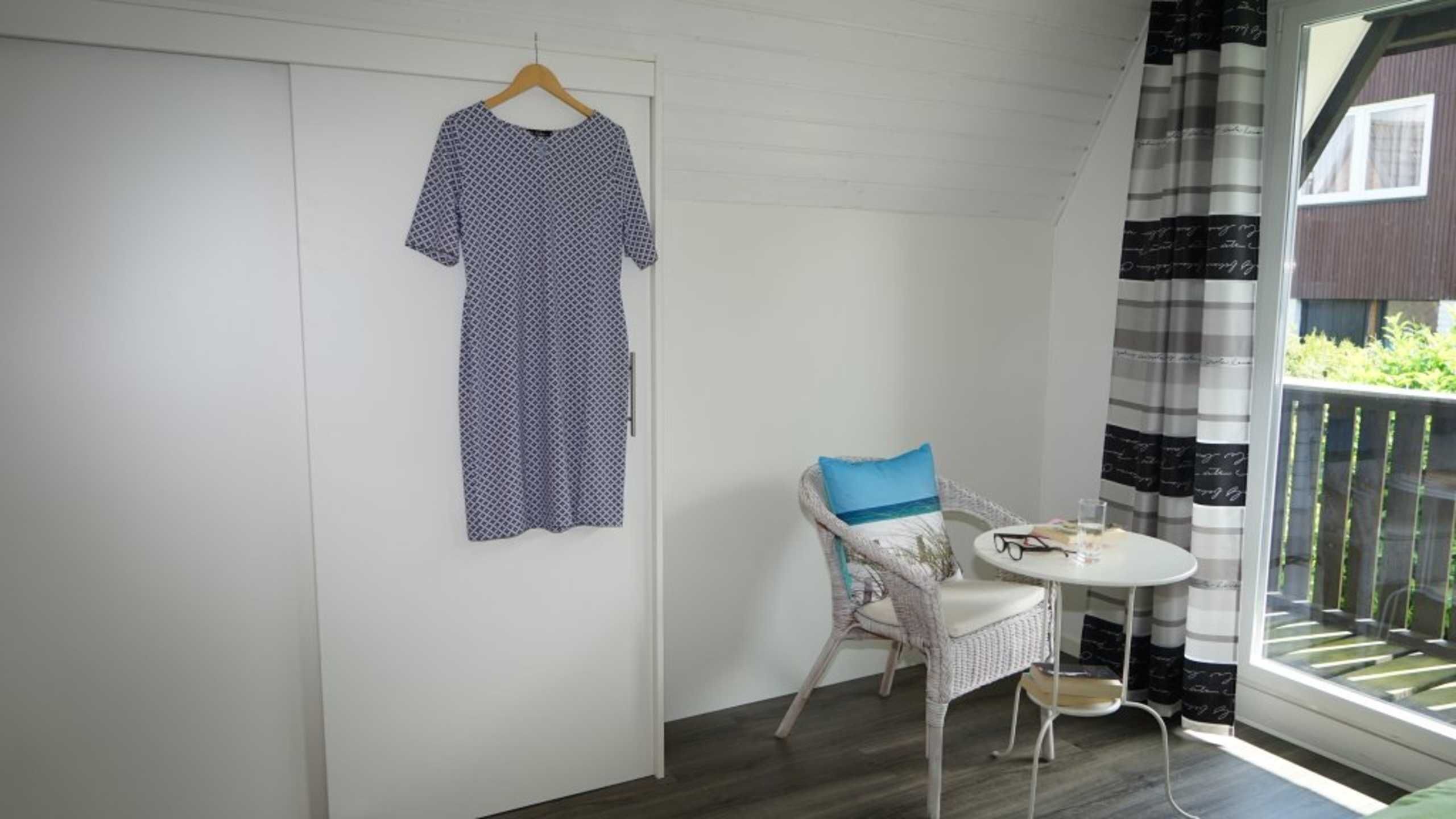 Habitació amb paret blanca, un vestit penjat, una cadira de vímet amb coixí blau i una taula rodona petita amb un got d'aigua i un llibre.