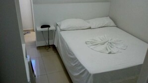 Ein kleines Zimmer mit einem Einzelbett, einer weißen Bettdecke, einem Nachttischtelefon und einem ordentlich gefalteten Handtuch auf dem Bett.