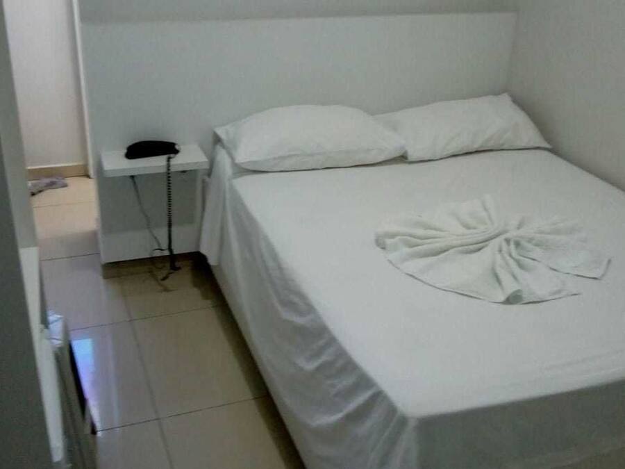 Una habitación pequeña con una cama individual, una colcha blanca, un teléfono en la mesita de noche y una toalla doblada con cuidado sobre la cama.