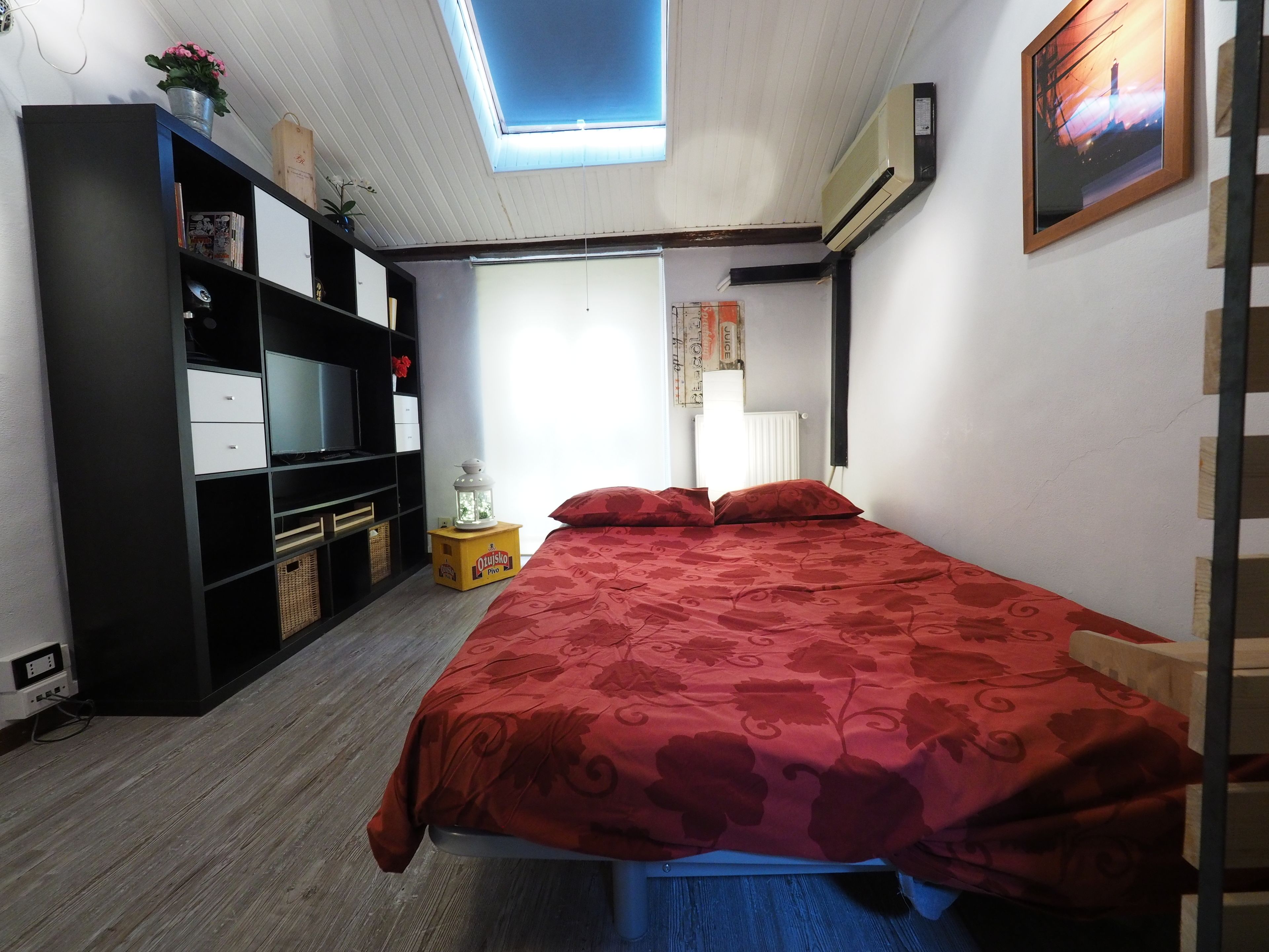 Ein Schlafzimmer mit Bett, Fernseher, Bücherregal, Heizkörper und Dachfenster.